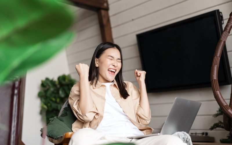 Joyful asian woman winning online on laptop, celebrating victory, shouting and rejoicing, triumphing.